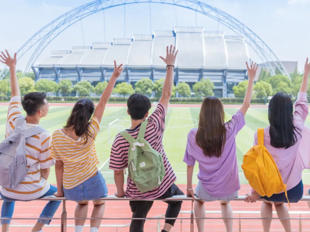 นักศึกษานั่งริมสนามกีฬามหาวิทยาลัย ยกมือขึ้นพร้อมกันในบรรยากาศสดใส