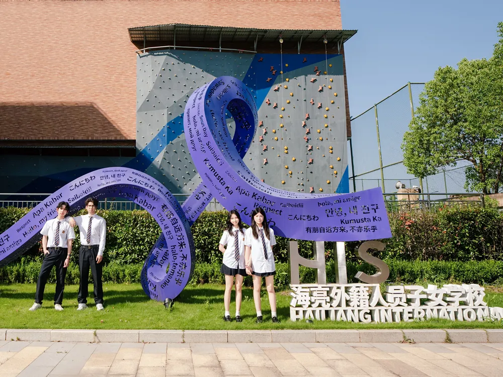 นักเรียนยืนหน้าสัญลักษณ์ HIS Haijiang International School พร้อมประติมากรรมคำทักทายหลายภาษา