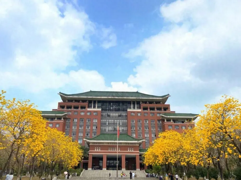 อาคารเรียนหลักของ South China Agricultural University สถาปัตยกรรมจีนร่วมสมัย หลังคากระเบื้องสีเขียว บันไดทางขึ้นกว้าง รายล้อมด้วยต้นไม้ดอกสีเหลืองและเสาธงแดงหน้ามหาวิทยาลัย