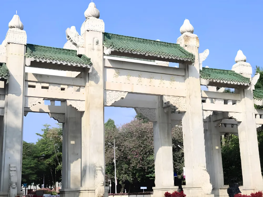 ซุ้มประตูหินสถาปัตยกรรมจีนดั้งเดิมของ South China Agricultural University หลังคากระเบื้องสีเขียว ประดับลวดลายแกะสลัก รายล้อมด้วยต้นไม้และท้องฟ้าแจ่มใส