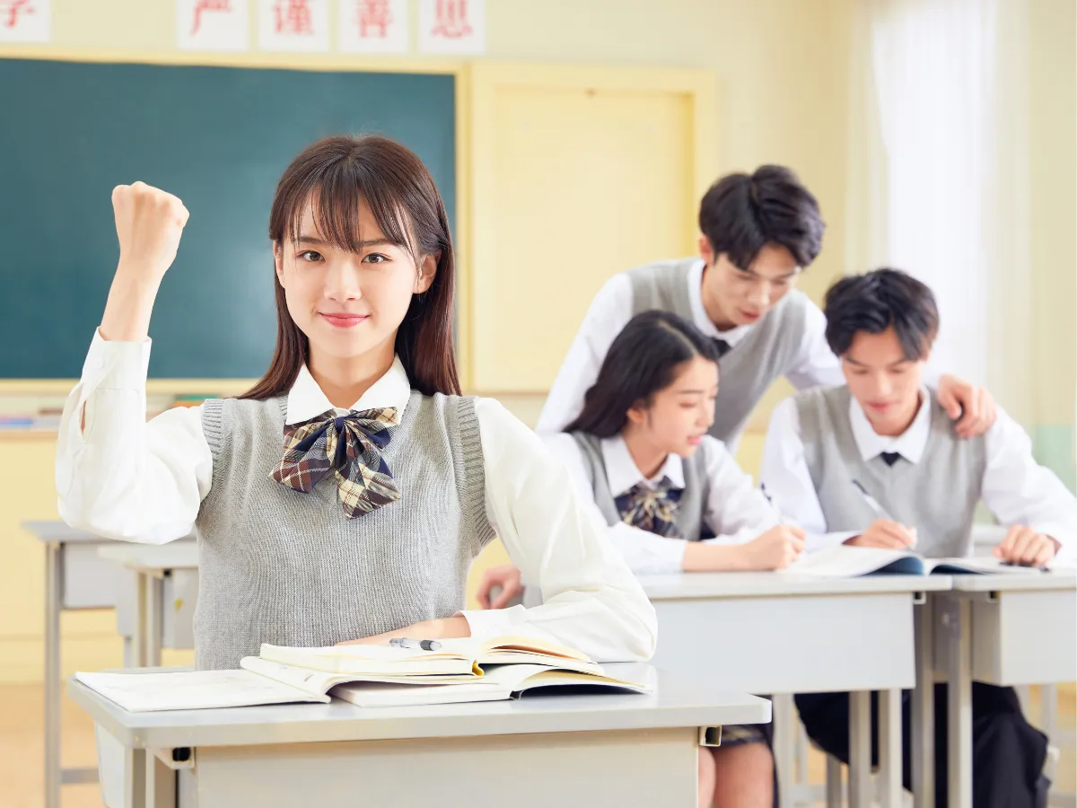นักเรียนหญิงในชุดเครื่องแบบนักเรียนมัธยมต้นหน้าตาน่ารัก กำลังนั่งอยู่ที่โต๊ะเรียนและชูหมัดขวาขึ้นด้วยท่าทางมั่นใจในตัวเอง โดยมีเพื่อน ๆ ในกลุ่มกำลังตั้งใจอ่านและเขียนหนังสืออยู่ด้านหลัง
